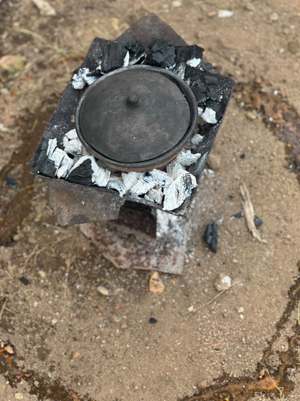 A covered earthen pot resting among the coals